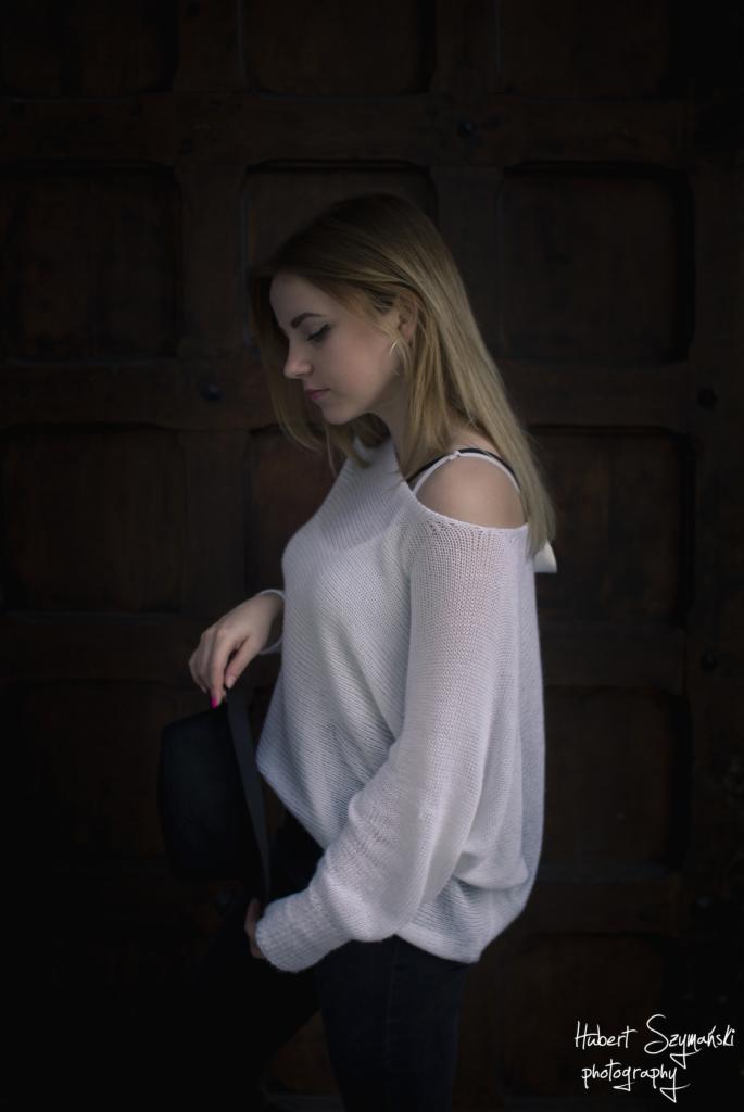 Portret młodej kobiety w swetrze z odkrytym ramieniem, trzymającej czarny kapelusz na tle ciemnych, drewnianych drzwi.