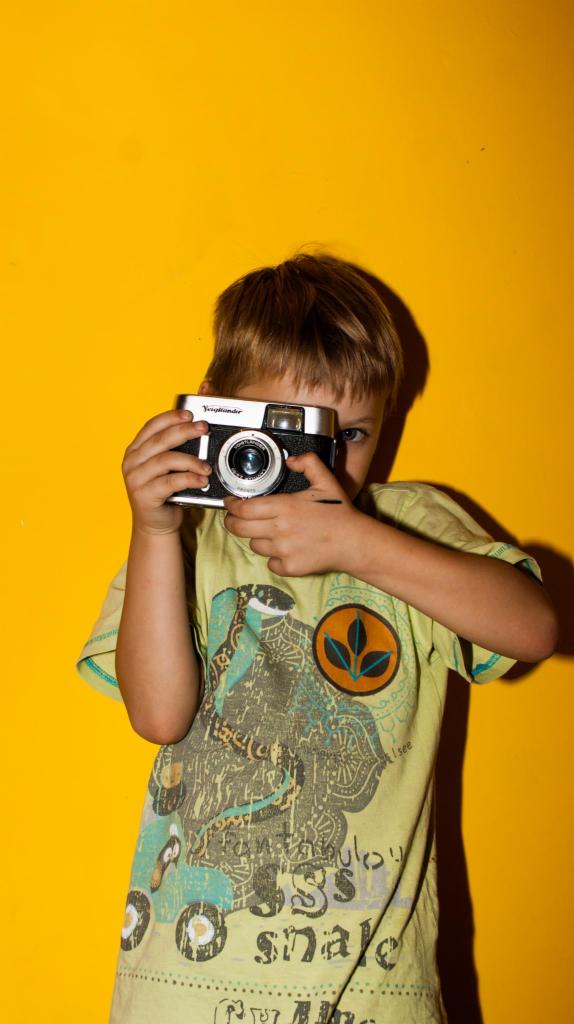 Chłopiec celuje w obiektyw aparatem Voigtländer na tle żółtej ściany, ubrany w koszulkę z nadrukiem węża.