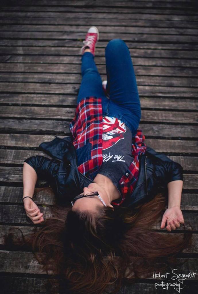 Młoda kobieta leżąca na drewnianym pomoście, ubrana w jeansy, koszulkę z nadrukiem, flanelową koszulę i skórzaną kurtkę, w czerwonych trampkach, z głową skierowaną w dół kadru.