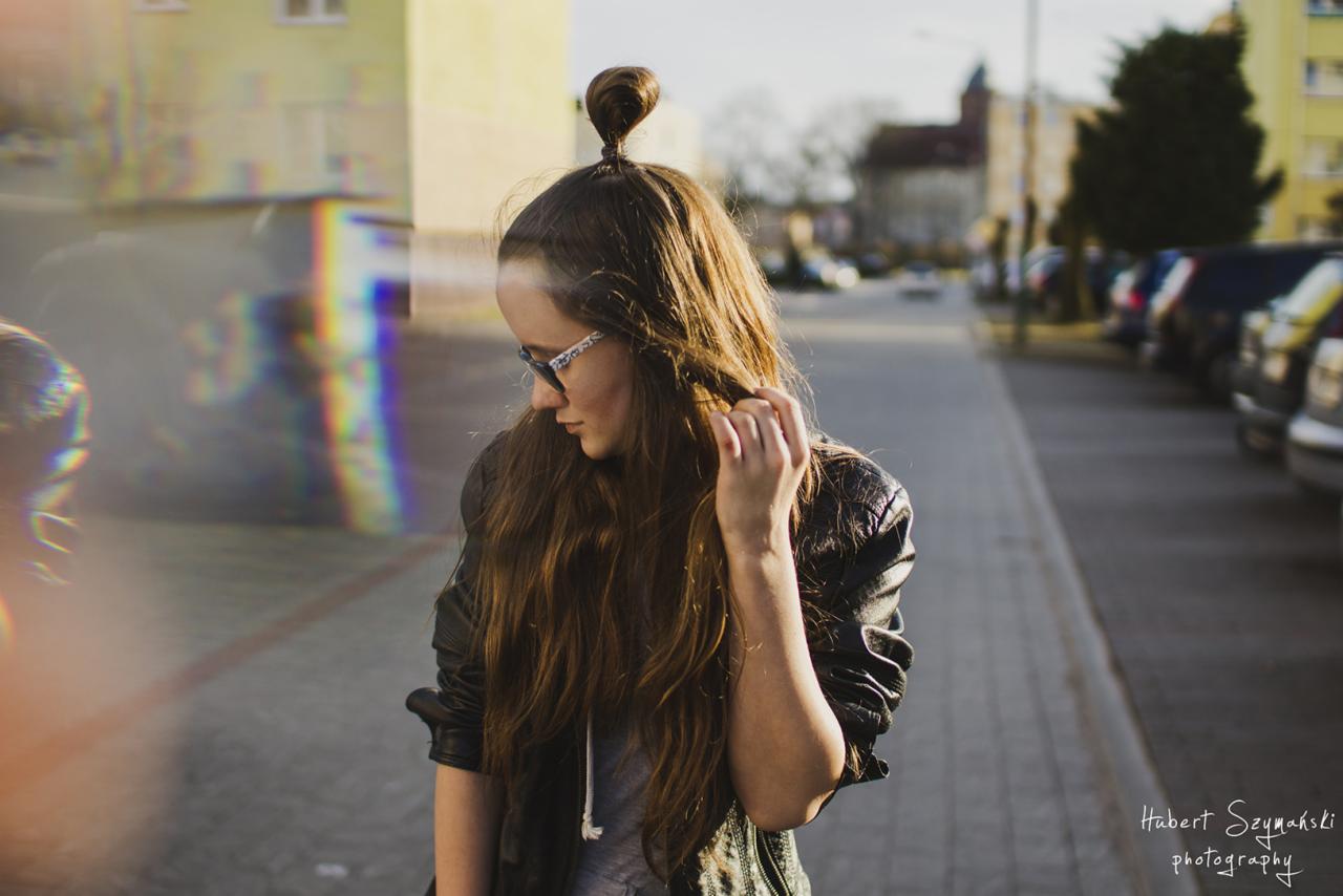 Portret młodej kobiety z długimi brązowymi włosami upiętymi w mały kok na czubku głowy, w skórzanej kurtce i okularach w ozdobnej oprawce, na tle miejskiej ulicy z budynkami i samochodami.