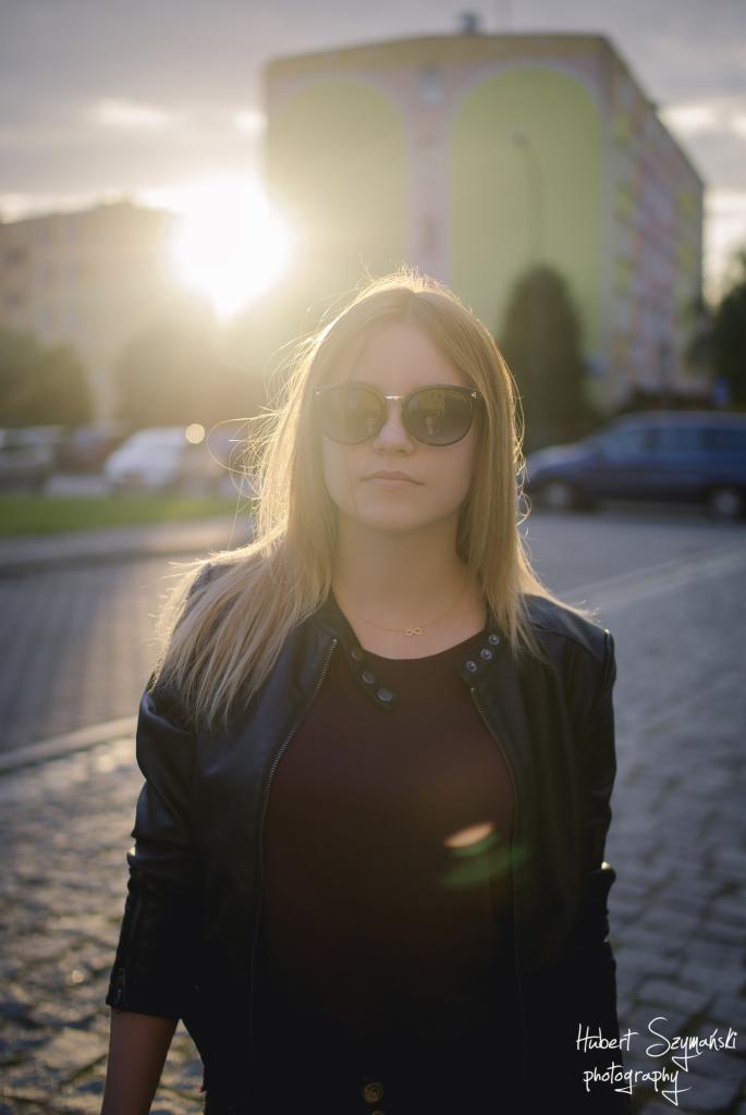 Portret kobiety w okularach przeciwsłonecznych na tle miejskiego krajobrazu w promieniach zachodzącego słońca, z widocznym budynkiem i brukowaną ulicą.