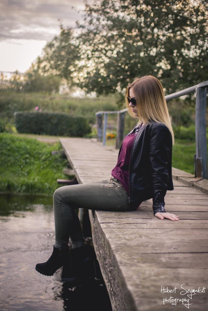 Kobieta w okularach przeciwsłonecznych, skórzanej kurtce i oliwkowych spodniach siedzi na drewnianym mostku nad rzeką, zanurzając stopę w wodzie; w tle drzewa i zachmurzone niebo.