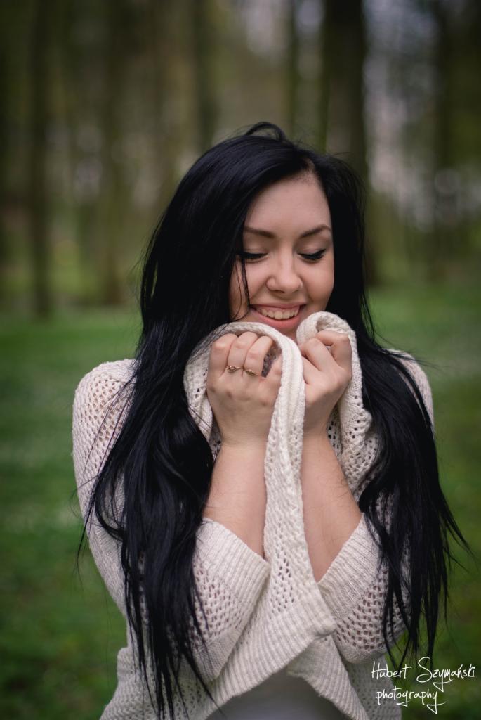 Portret uśmiechniętej brunetki w jasnym swetrze, trzymającej biały szal przy twarzy, na tle rozmytego leśnego krajobrazu.