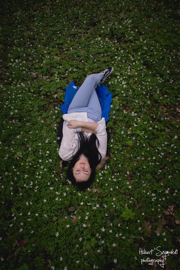 Młoda kobieta leżąca na łące pełnej białych kwiatów, ubrana w jasne jeansy i sweter, z zamkniętymi oczami, trzymająca książkę, z widocznym logo fotografa w prawym dolnym rogu.