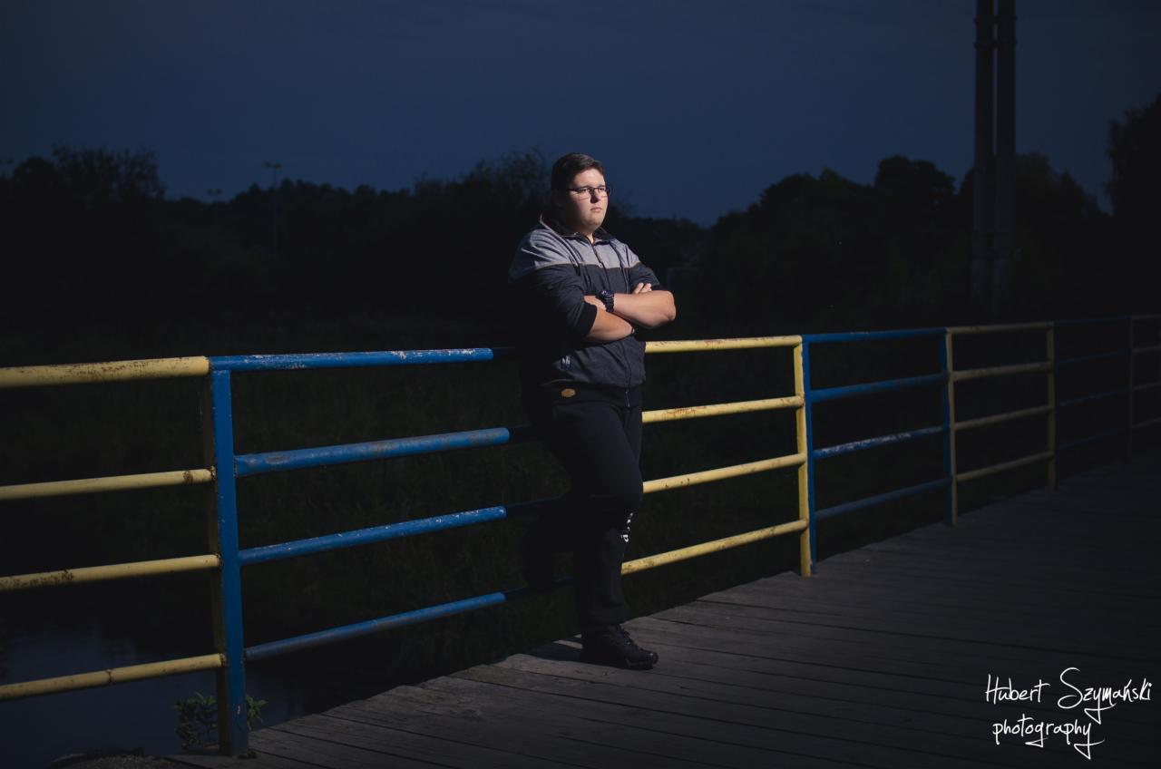 Mężczyzna w okularach, oparty o barierki mostu pomalowane na niebiesko i żółto, w ciemnym otoczeniu z widocznym ciemnym tłem roślinności.