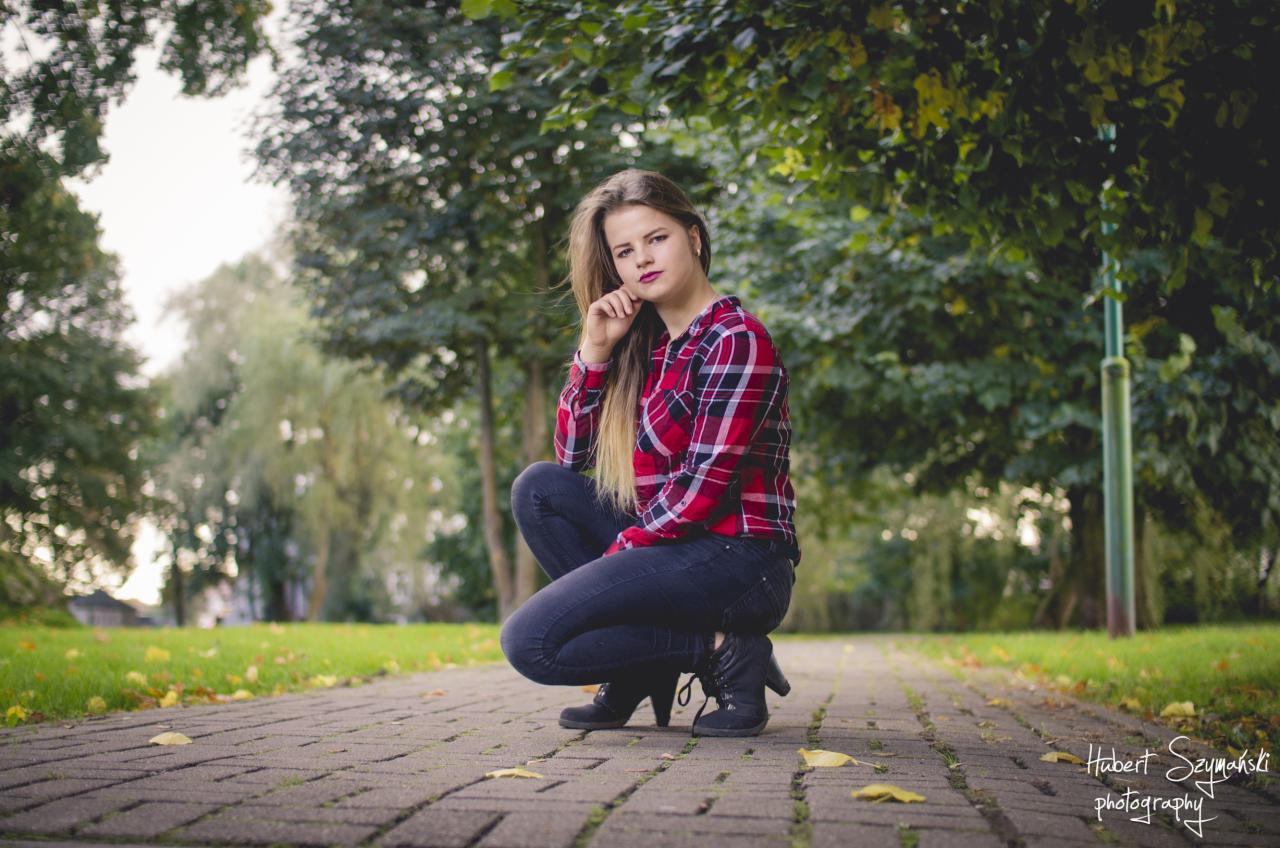 Młoda kobieta w czerwonej koszuli w kratę kuca na brukowanej ścieżce w parku, z drzewami w tle, pozując do zdjęcia.