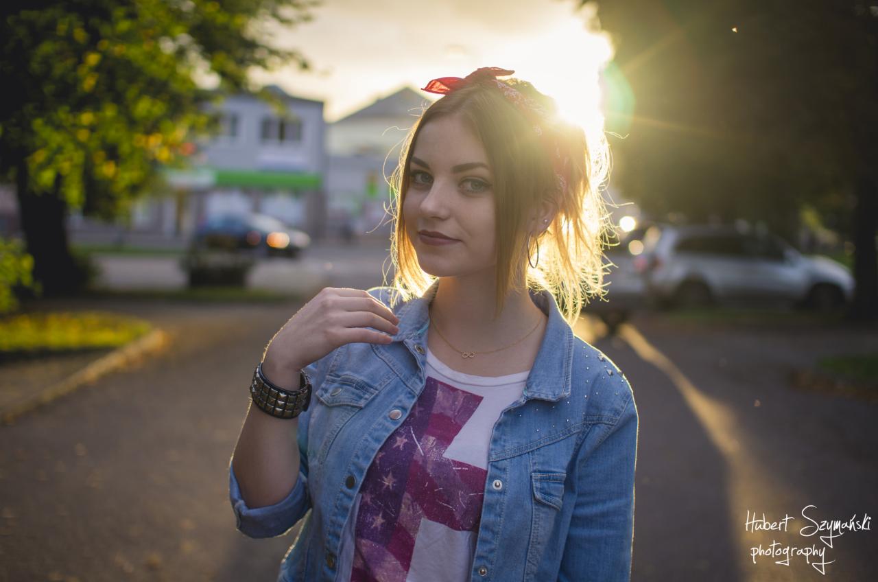 Portret młodej kobiety w dżinsowej kurtce, z czerwoną chustą we włosach, uchwycony pod słońce na tle ulicy i budynków. Złota biżuteria na szyi i nadgarstku, delikatny uśmiech.