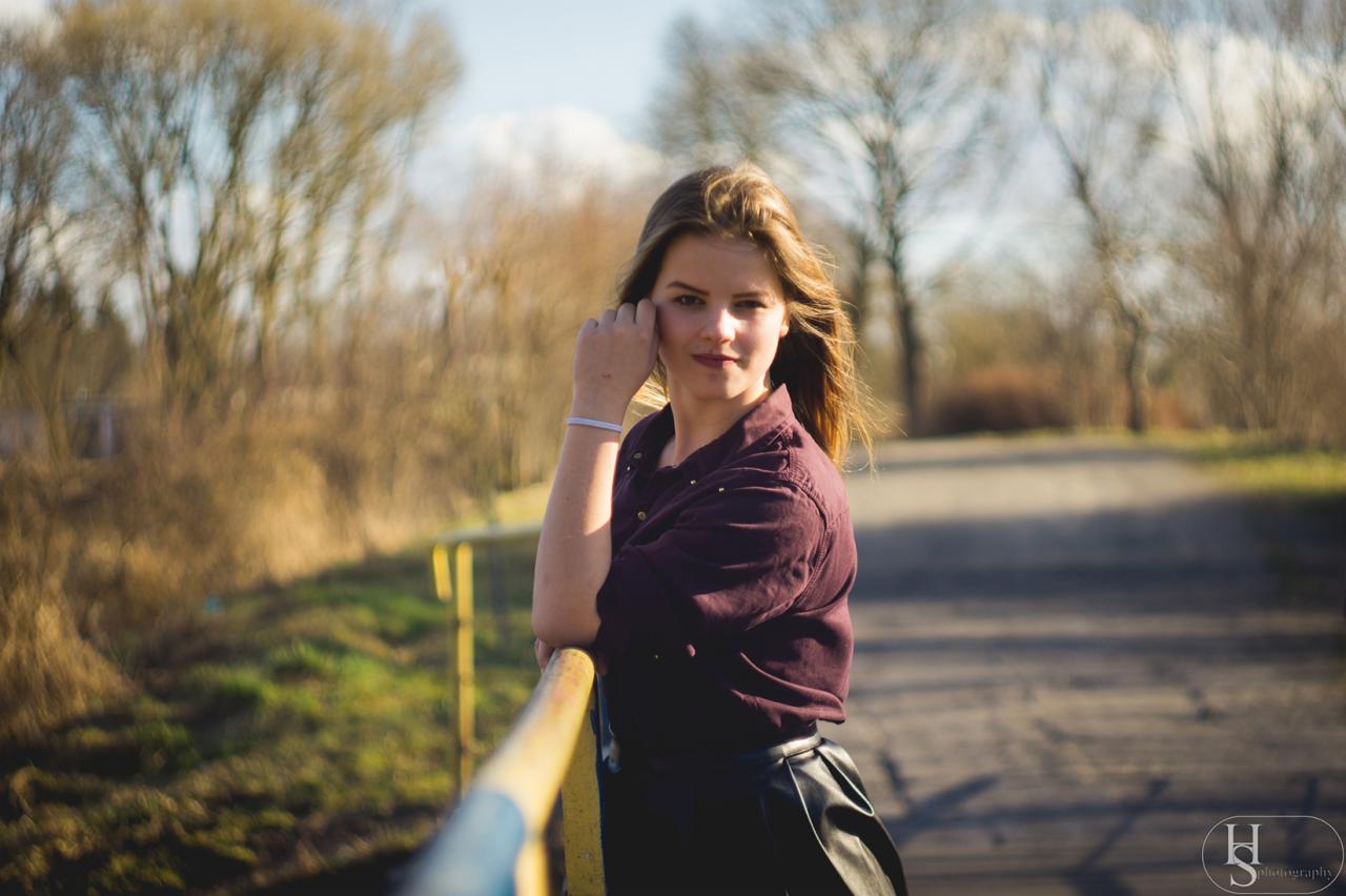 Portret młodej kobiety w bordowej koszuli opierającej się o żółtą barierkę, z rozmyciem tła ukazującym drzewa i drogę.