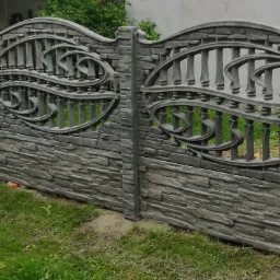 Szary betonowy płot z dekoracyjnym wzorem i imitacją kamienia, widok z boku na tle zieleni.