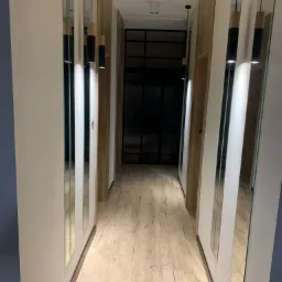 Wąski korytarz z drewnianą podłogą, białymi ścianami i lustrami w pionowych ramach, oświetlony trzema wiszącymi lampami z drewnianymi i czarnymi elementami.