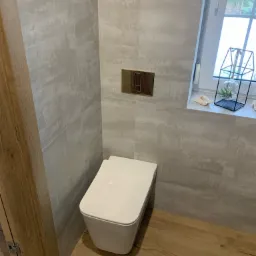 Nowoczesna łazienka z wiszącą toaletą, szarymi płytkami imitującymi beton i drewnopodobną podłogą, uzupełniona o dekoracje okienne.