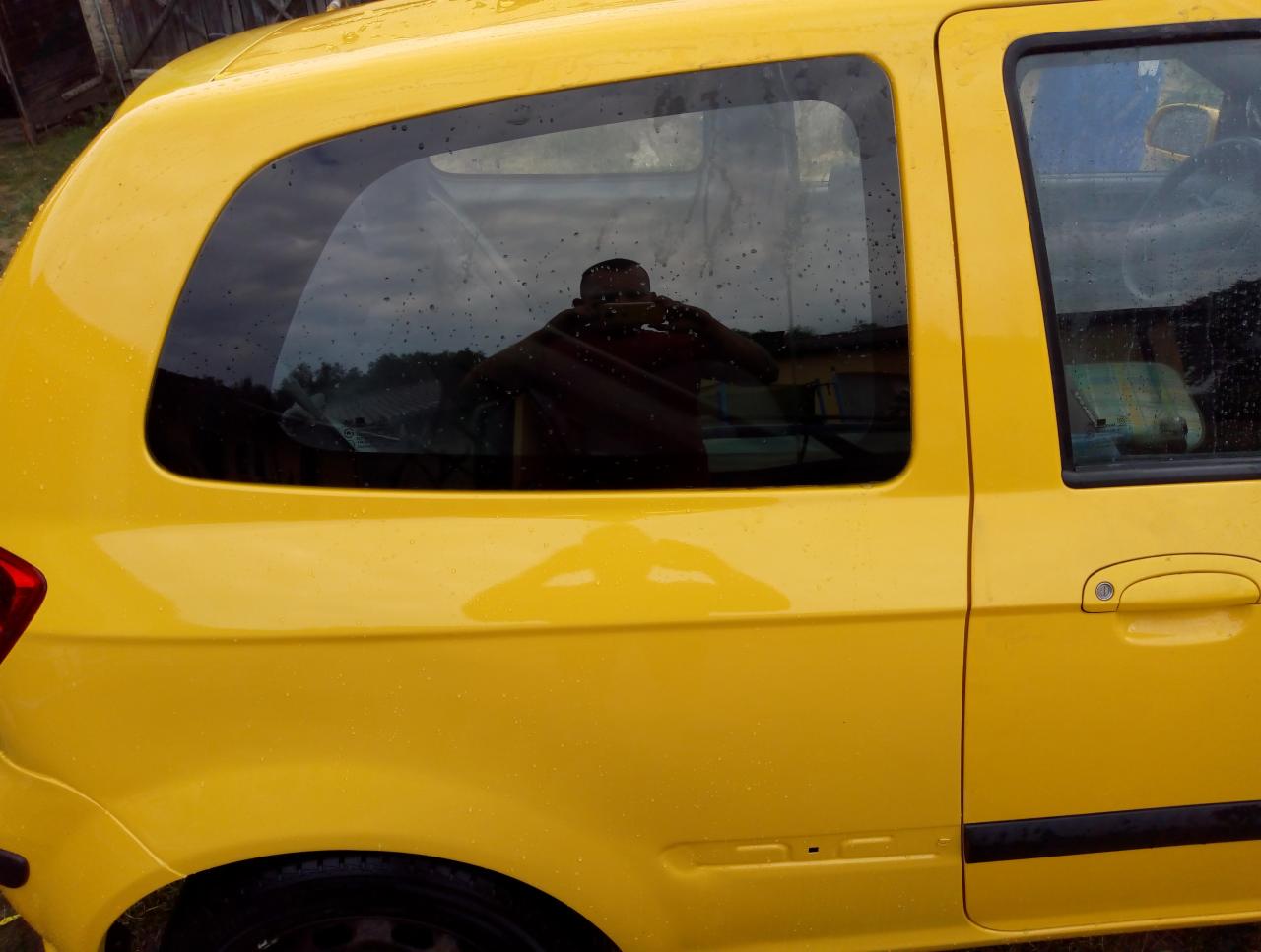 Żółty samochód po myciu, widoczne krople wody na karoserii i odbicie osoby w szybie.