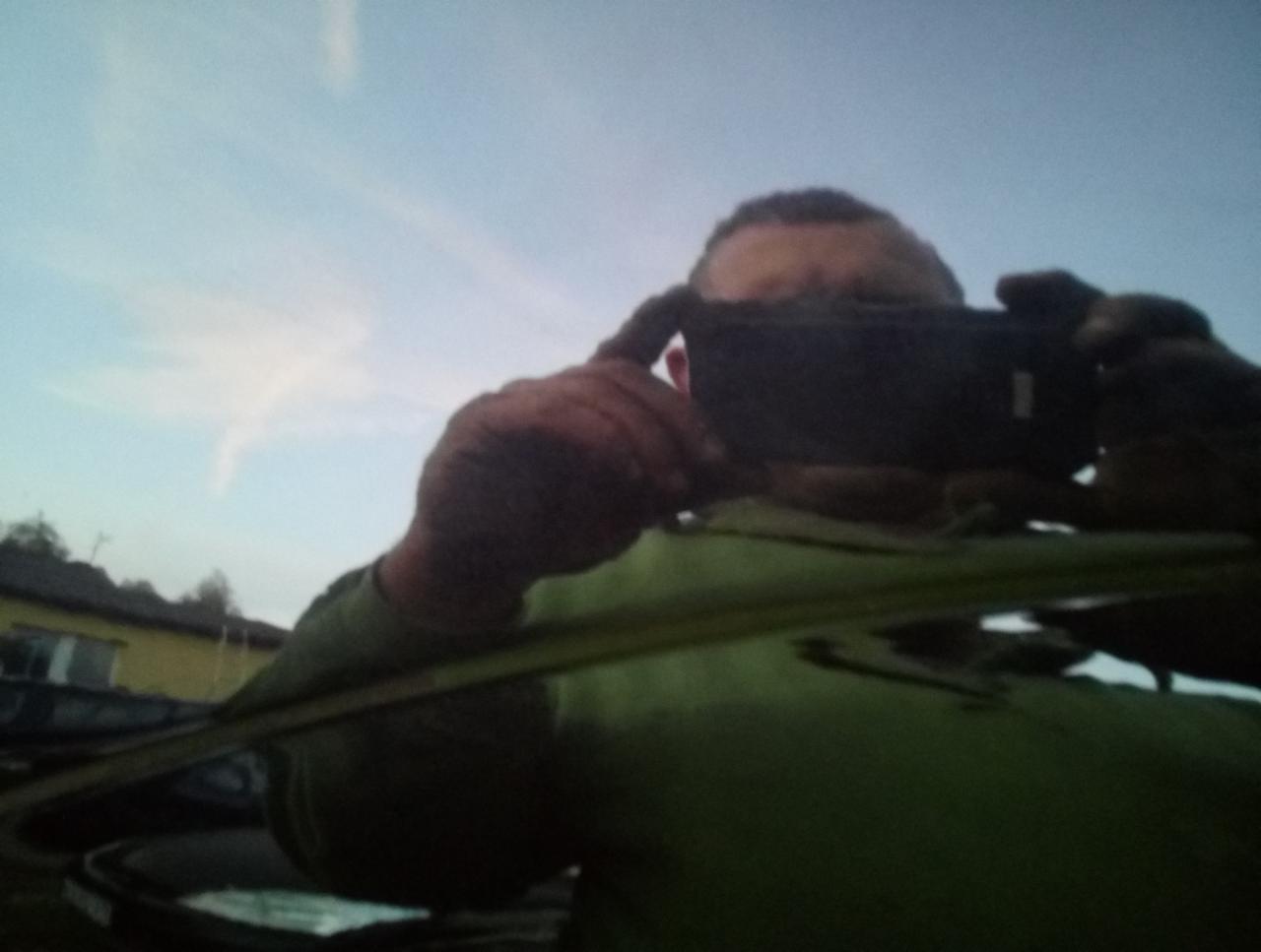 Mężczyzna robi zdjęcie smartfonem odbicia w lakierowanej powierzchni samochodu, widoczne niebo i fragment budynku.