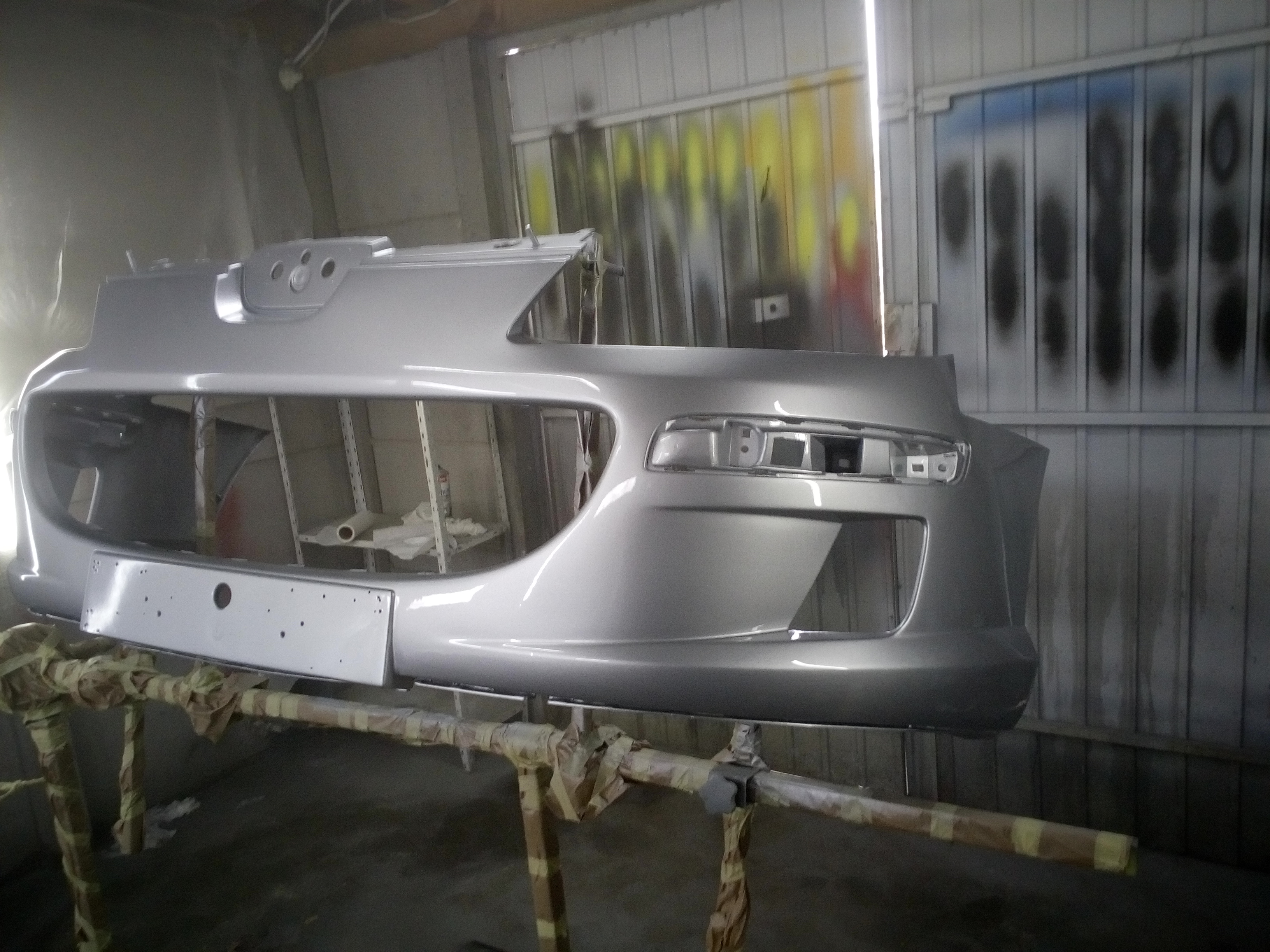 Pomalowany na srebrno zderzak samochodowy na stojaku w warsztacie, widoczne detale mocowania tablicy rejestracyjnej i ślady po lakierowaniu na ścianach.