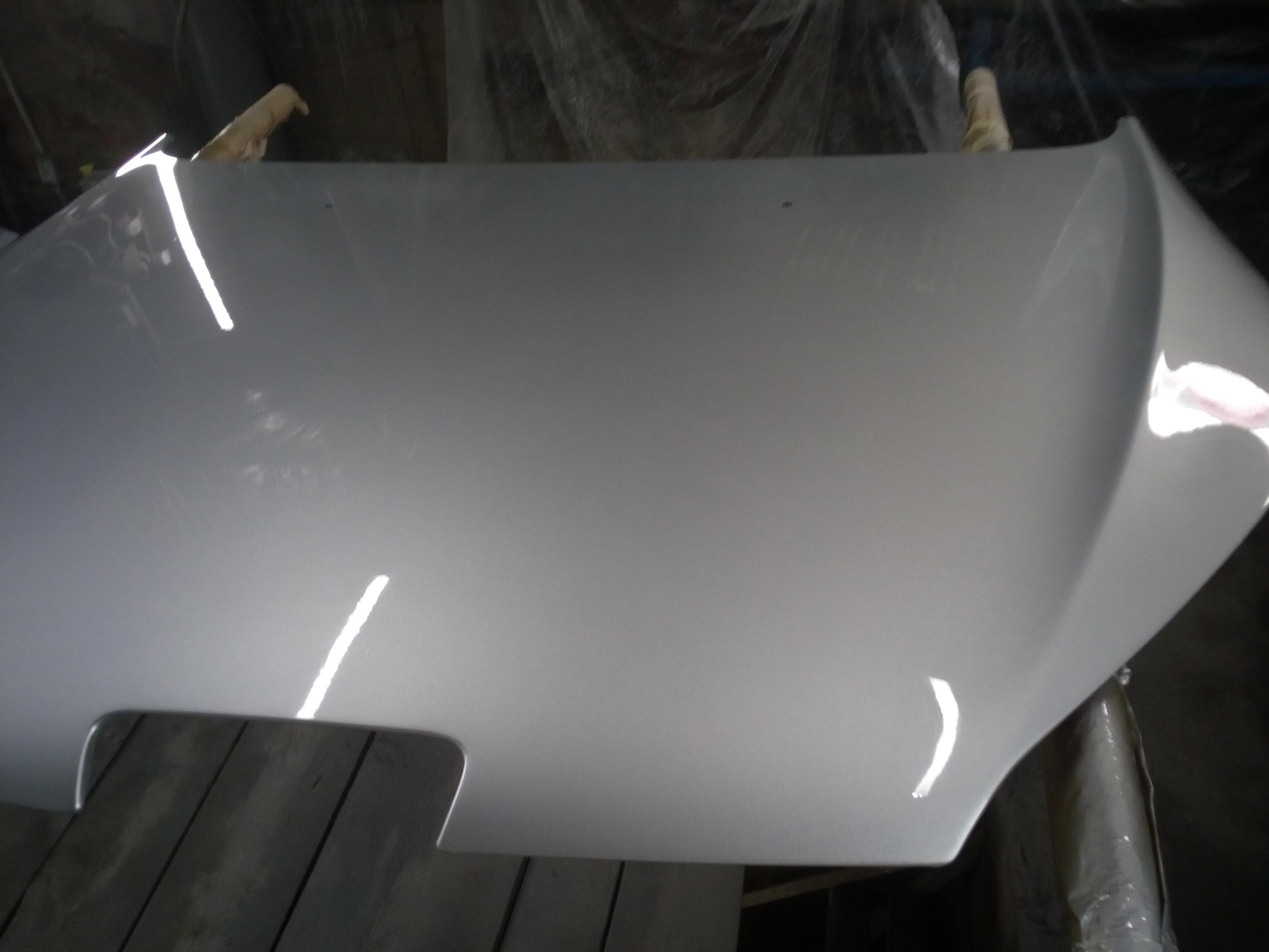 Świeżo polakierowana, srebrna maska samochodu, leżąca na drewnianym stole w warsztacie, z widocznymi odbiciami światła na gładkiej powierzchni.