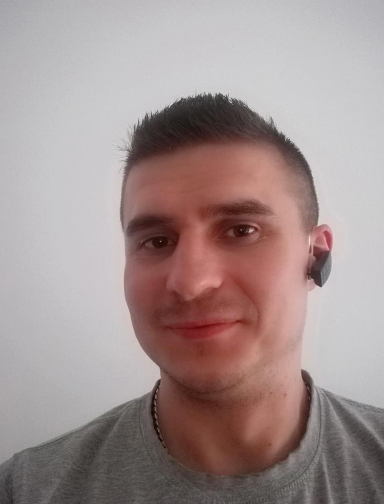 Mężczyzna z krótkimi włosami i czarnym urządzeniem w uchu, prawdopodobnie słuchawka lub aparat słuchowy, na tle białej ściany.