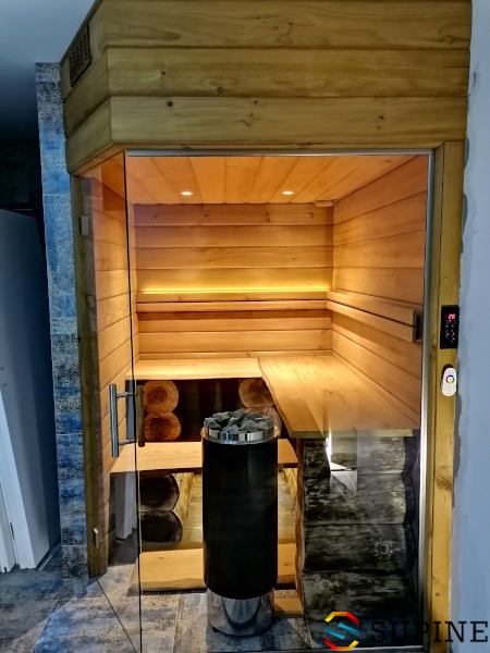 Wnętrze sauny z piecem na kamienie, drewnianymi ławkami i szklanymi drzwiami; widoczne oświetlenie punktowe i panel sterowania temperaturą.