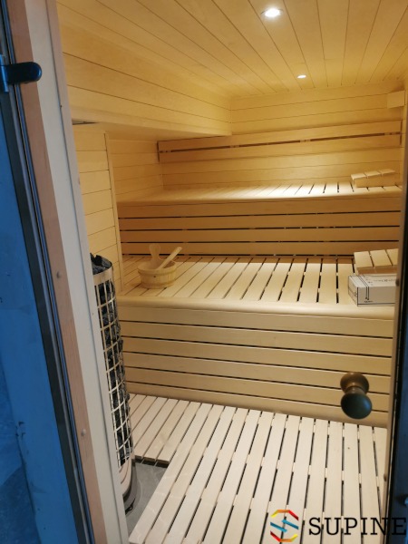 Wnętrze nowej, jasnej sauny z drewnianymi ławkami, piecem elektrycznym i akcesoriami do aromaterapii, widoczne przez uchylone drzwi.