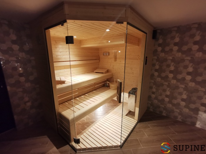 Wnętrze narożnej kabiny z jasnego drewna, przeszklonej z dwóch stron, z dwoma poziomami ławek i piecem na kamienie, widoczne z zewnątrz na tle ściany wyłożonej mozaiką.