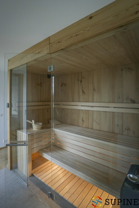 Wnętrze nowoczesnej drewnianej kabiny z przeszklonymi drzwiami, widoczne ławy, wiaderko i piec.