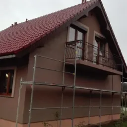 Dom jednorodzinny z ocieploną elewacją w trakcie prac wykończeniowych, widoczne rusztowania i fragmenty niezakończonych prac tynkarskich.