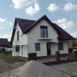 Świeżo otynkowany dom jednorodzinny z brązowym dachem i ceglanym ogrodzeniem w trakcie budowy, widoczne rusztowania i hałda piasku na pierwszym planie.