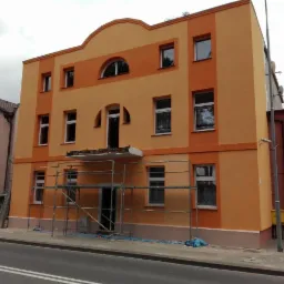 Dwukolorowa elewacja budynku po remoncie, z widocznymi rusztowaniami na pierwszym planie i świeżo pomalowaną fasadą w odcieniach pomarańczy.