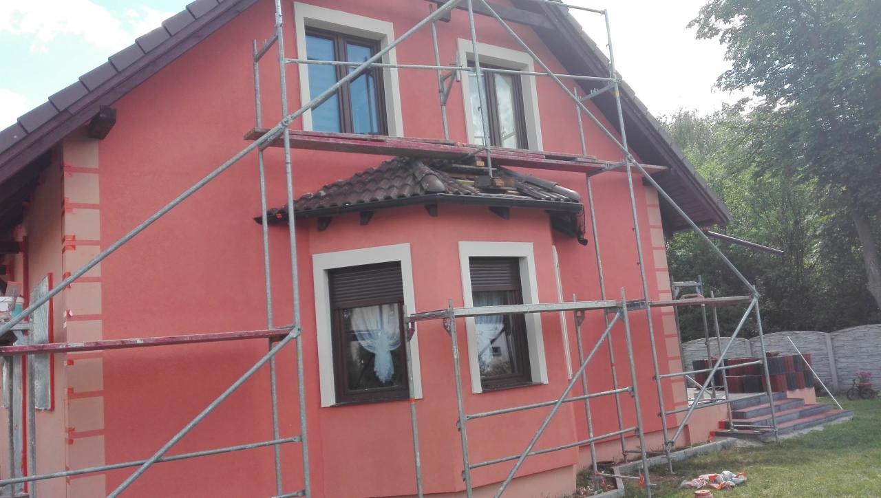 Dom jednorodzinny w trakcie prac elewacyjnych, widoczne rusztowania, fragmenty ocieplenia i świeżo nałożony tynk w odcieniach różu, z elementami dekoracyjnymi imitującymi cegłę.