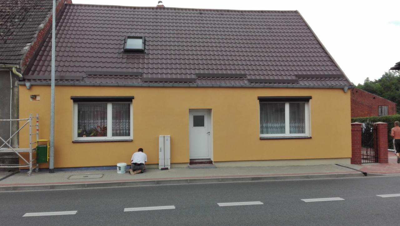 Żółta elewacja domu jednorodzinnego podczas malowania; widoczny pracownik klęczący przy ścianie z puszką farby, obok biała skrzynka elektryczna i fragment rusztowania.