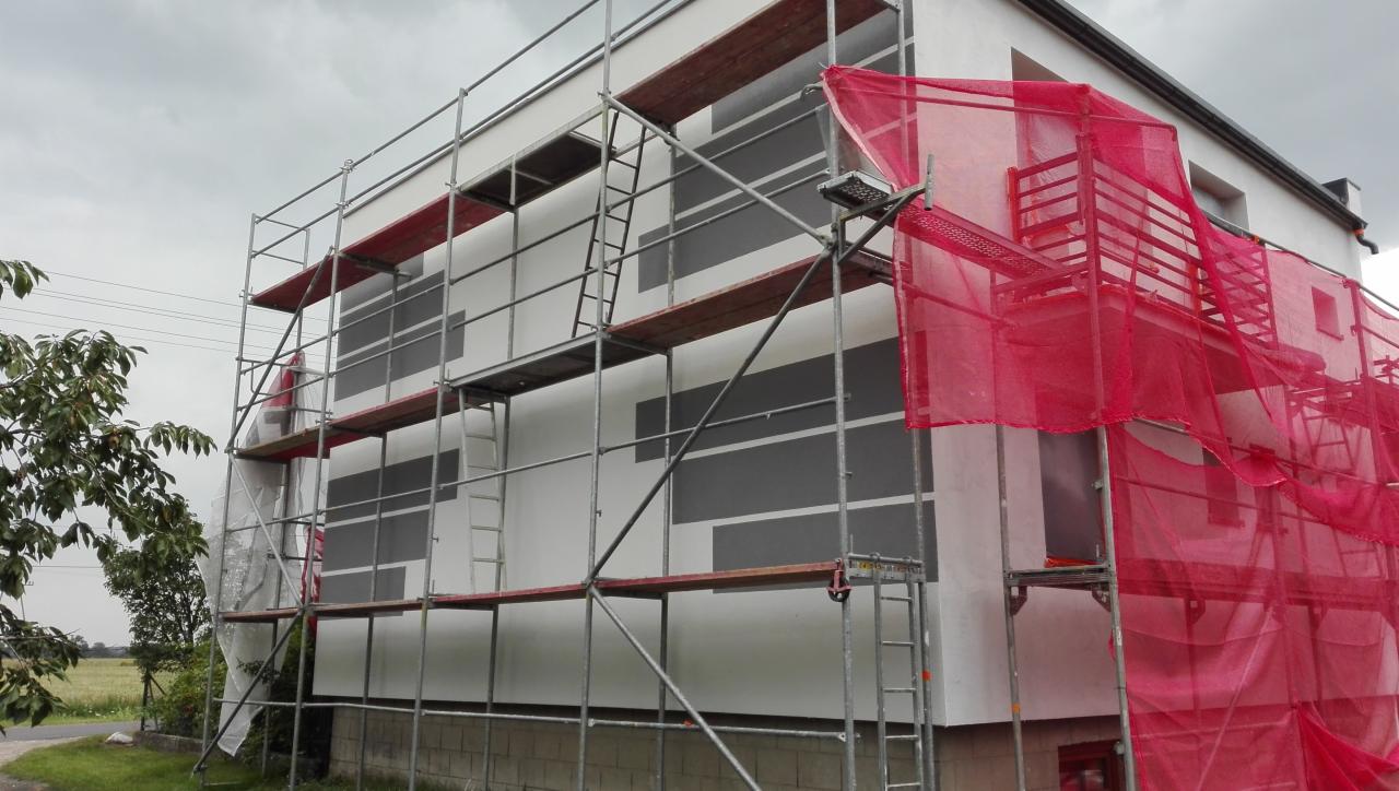 Dom w Mirosławcu w trakcie prac elewacyjnych, z widocznymi rusztowaniami, częściowo zakryty siatką ochronną, z fragmentami pomalowanej ściany w szare pasy.