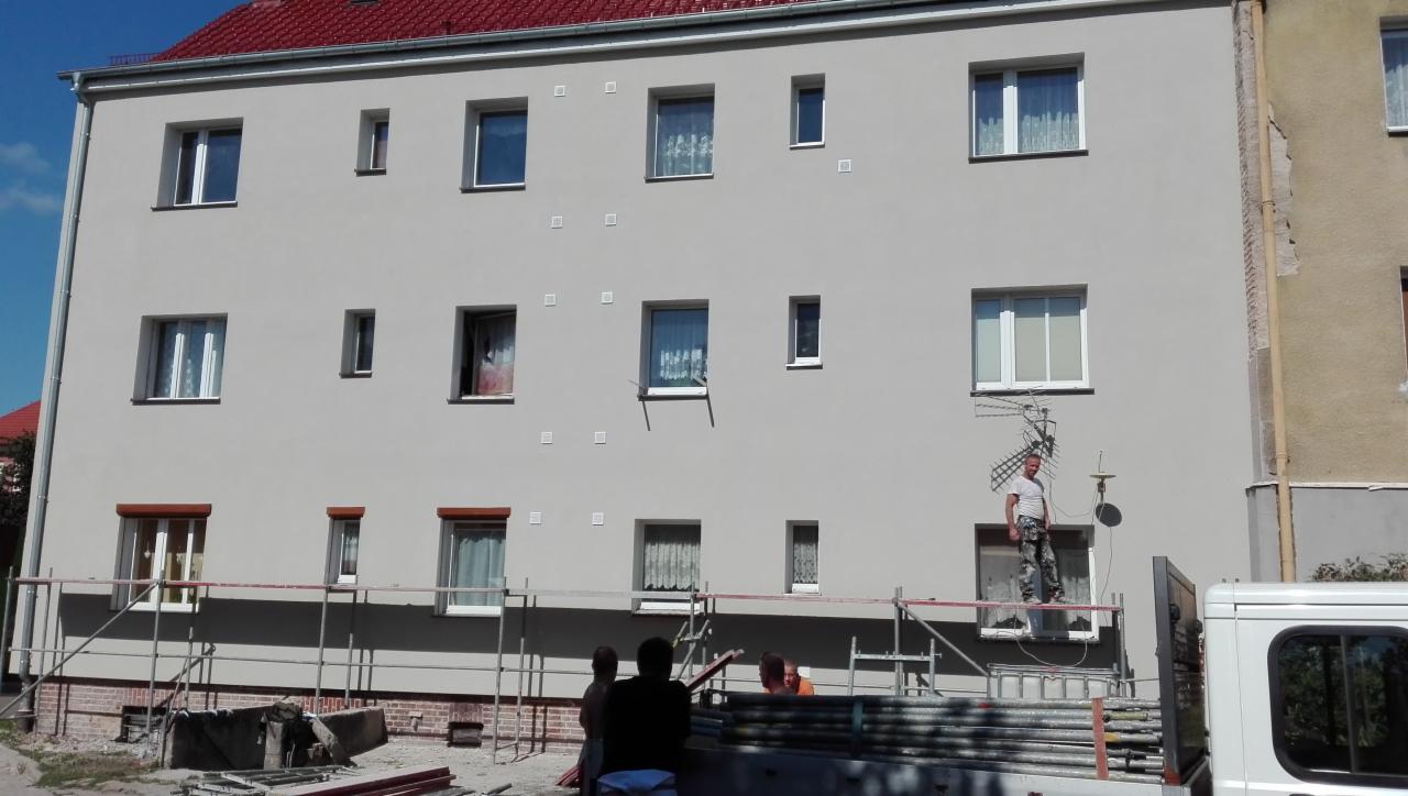 Pracownik na rusztowaniu podczas tynkowania elewacji budynku wielorodzinnego, widoczny fragment nieotynkowanej ściany i antena satelitarna.