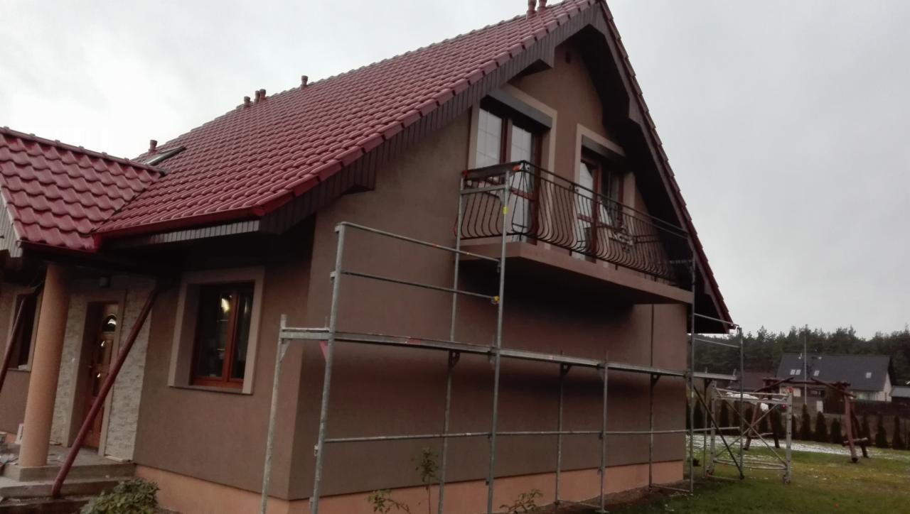 Dom jednorodzinny z ocieploną elewacją w trakcie prac wykończeniowych, widoczne rusztowania i fragmenty niezakończonych prac tynkarskich.