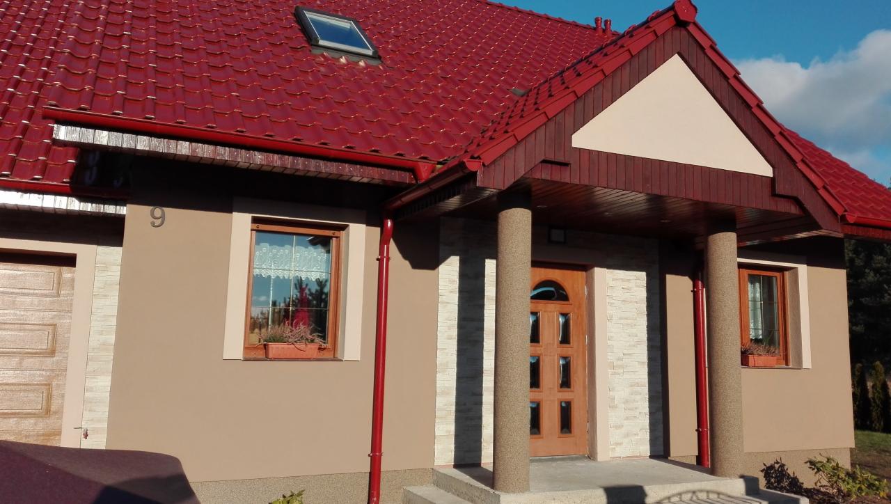 Dom jednorodzinny z czerwoną dachówką, beżową elewacją, oknami z kwiatami i numerem 9, widziany z niskiej perspektywy.