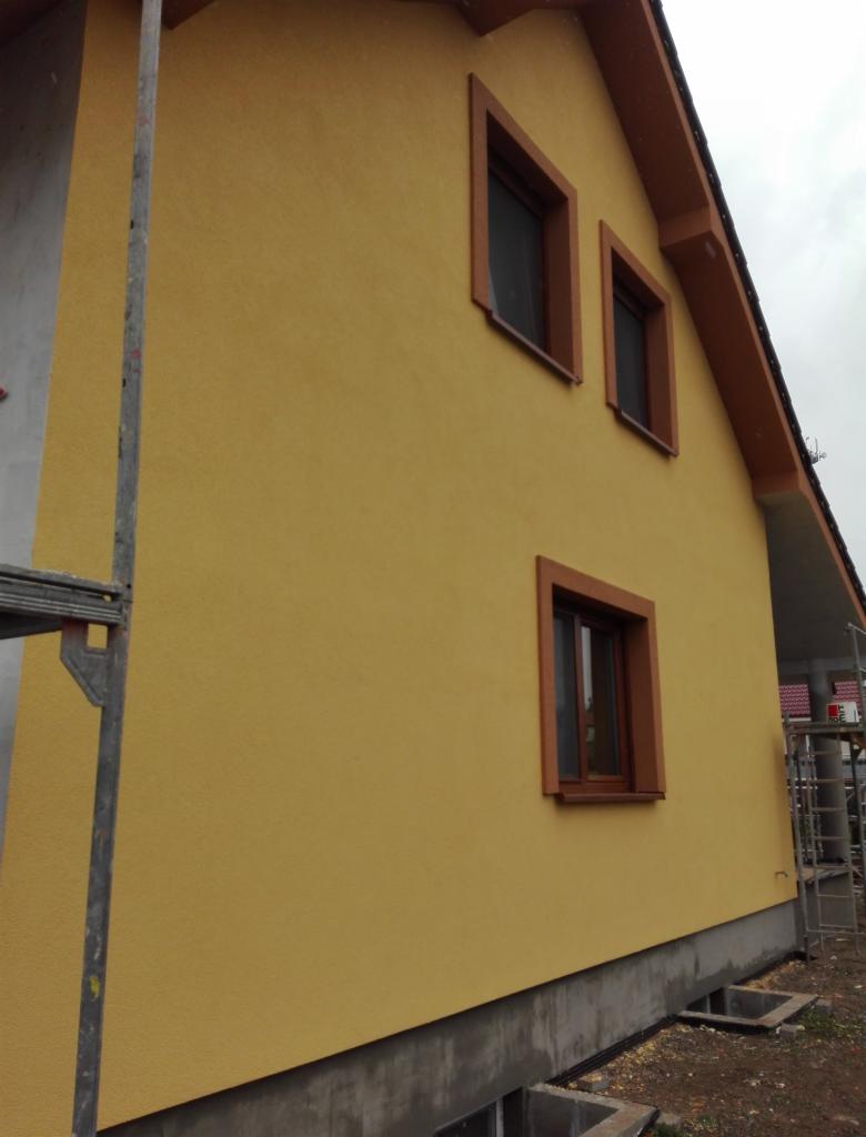 Żółta elewacja domu jednorodzinnego z brązowymi ramami okiennymi, widok z boku na tle pochmurnego nieba, z widocznymi fragmentami rusztowania.