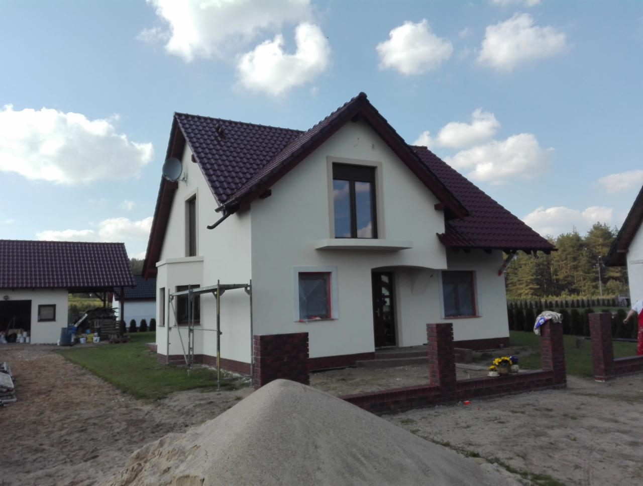 Świeżo otynkowany dom jednorodzinny z brązowym dachem i ceglanym ogrodzeniem w trakcie budowy, widoczne rusztowania i hałda piasku na pierwszym planie.