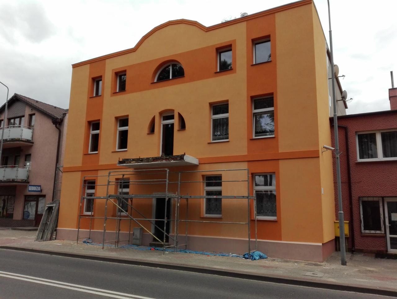 Dwukolorowa elewacja budynku po remoncie, z widocznymi rusztowaniami na pierwszym planie i świeżo pomalowaną fasadą w odcieniach pomarańczy.