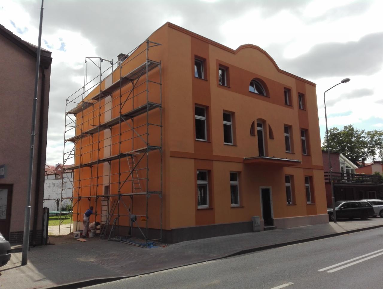 Dwukolorowa elewacja budynku w trakcie prac, z rusztowaniem i pracownikami malującymi dolną partię ściany na odcienie pomarańczy.