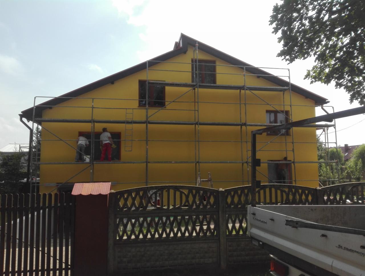 Żółty dom jednorodzinny w trakcie malowania elewacji, widoczne rusztowania, dwóch pracowników w trakcie pracy, jeden w czerwonych spodniach, ogrodzenie z przodu, przyczepka samochodowa w prawym...