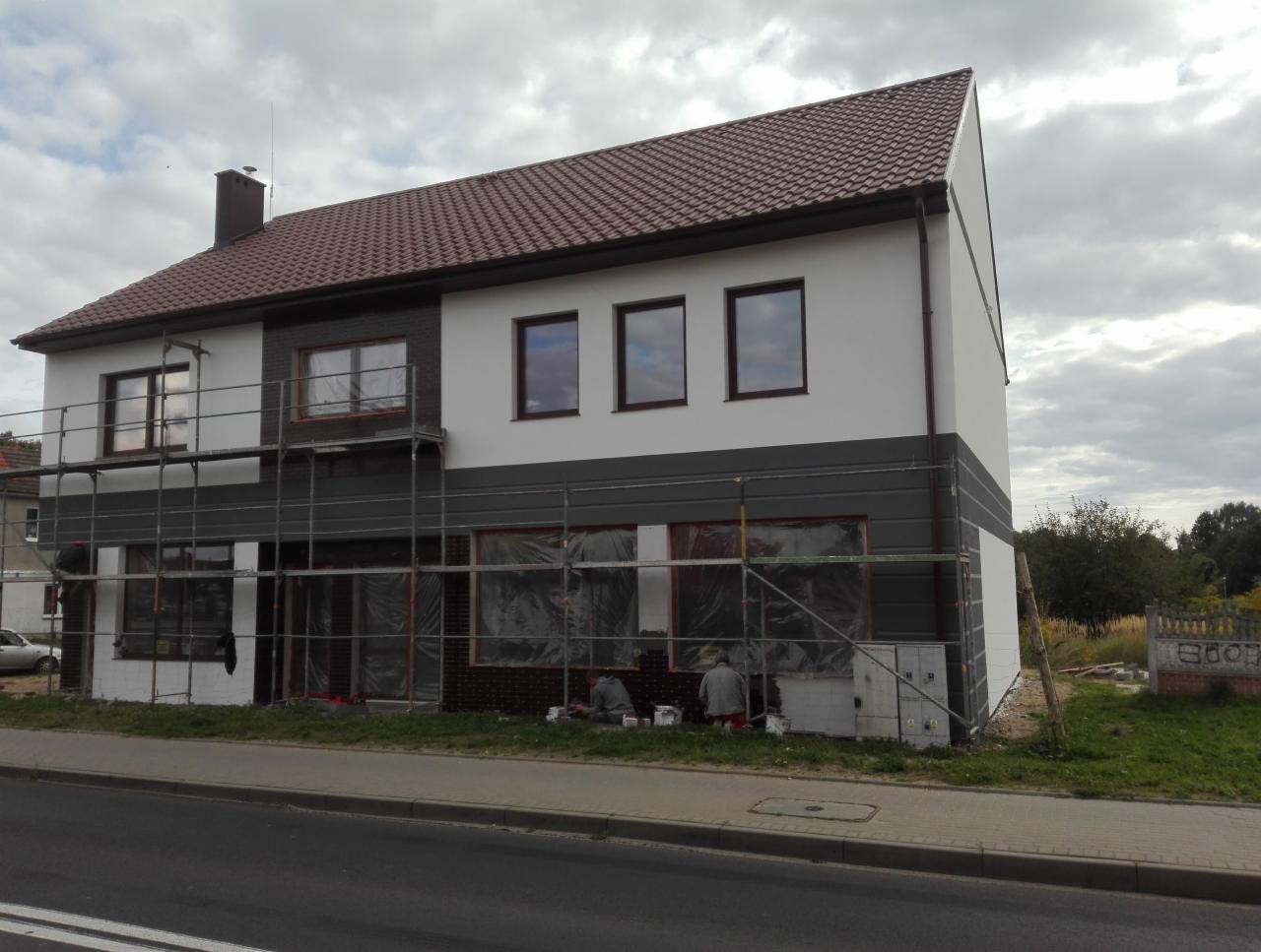 Dwukondygnacyjny dom w trakcie malowania elewacji, widoczne rusztowania, zabezpieczone okna folią i dwóch pracowników przy pracy na dole, szara kolorystyka dolnej części elewacji i biała górnej.