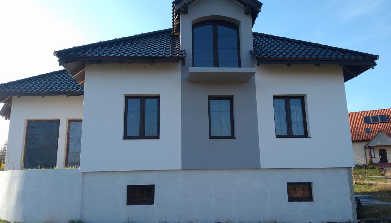 Nowoczesny dom jednorodzinny z jasną elewacją i ciemną dachówką, widok z boku, okna zabezpieczone folią w trakcie prac wykończeniowych.