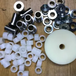 Różnorodne metalowe i plastikowe tuleje oraz biały krążek z otworem, ułożone na drewnianej powierzchni.