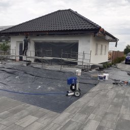 Usługi budowlane Wiesław Lisiecki - Pracownik na rusztowaniu maluje elewację domu natryskowo za pomocą agregatu malarskiego; zabezpieczona folią okna i teren wokół budynku.