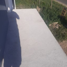 Usługi budowlane Wiesław Lisiecki - Świeżo otynkowana elewacja budynku z widocznym cieniem osoby robiącej zdjęcie, na tle zieleni i fragmentu drogi.