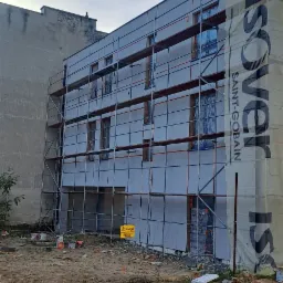 Budynek w trakcie ocieplania elewacji, widoczne rusztowania, częściowo zakryty folią z logo Isover Saint-Gobain, teren budowy z farbami i gruzem.