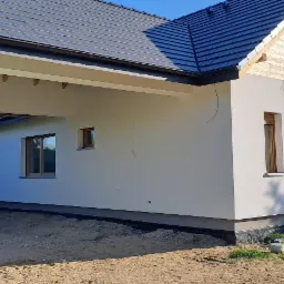 Nowoczesny dom jednorodzinny z jasną elewacją o fakturze baranka i drewnianymi oknami, widok z zewnątrz na świeżo otynkowaną ścianę i fragment dachu z grafitowej dachówki, z przodu widoczna...