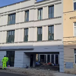 Nowoczesny budynek z jasną elewacją w trakcie wykańczania; pracownicy w jaskrawych kombinezonach malują dolną część elewacji, widoczne rusztowania i materiały budowlane.