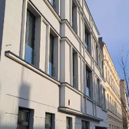 Pracownicy w trakcie malowania elewacji budynku z szarą fasadą, widoczne okna z szybami, drabina, wiadro z farbą i przechodnie w tle