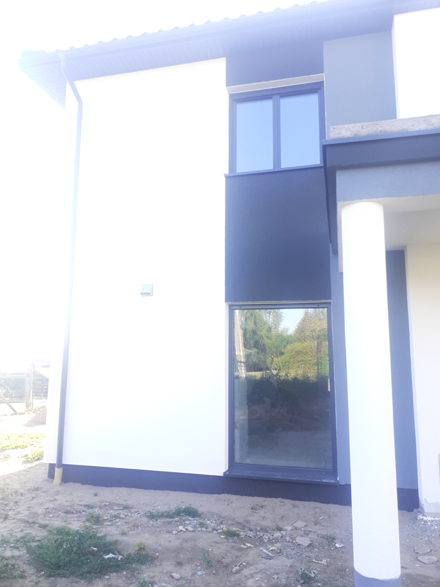 Nowoczesna, dwukondygnacyjna elewacja domu z białymi ścianami i grafitowymi wstawkami, widoczne okna z refleksami drzew, świeżo pomalowany cokół, teren wokół budynku jeszcze niezagospodarowany.