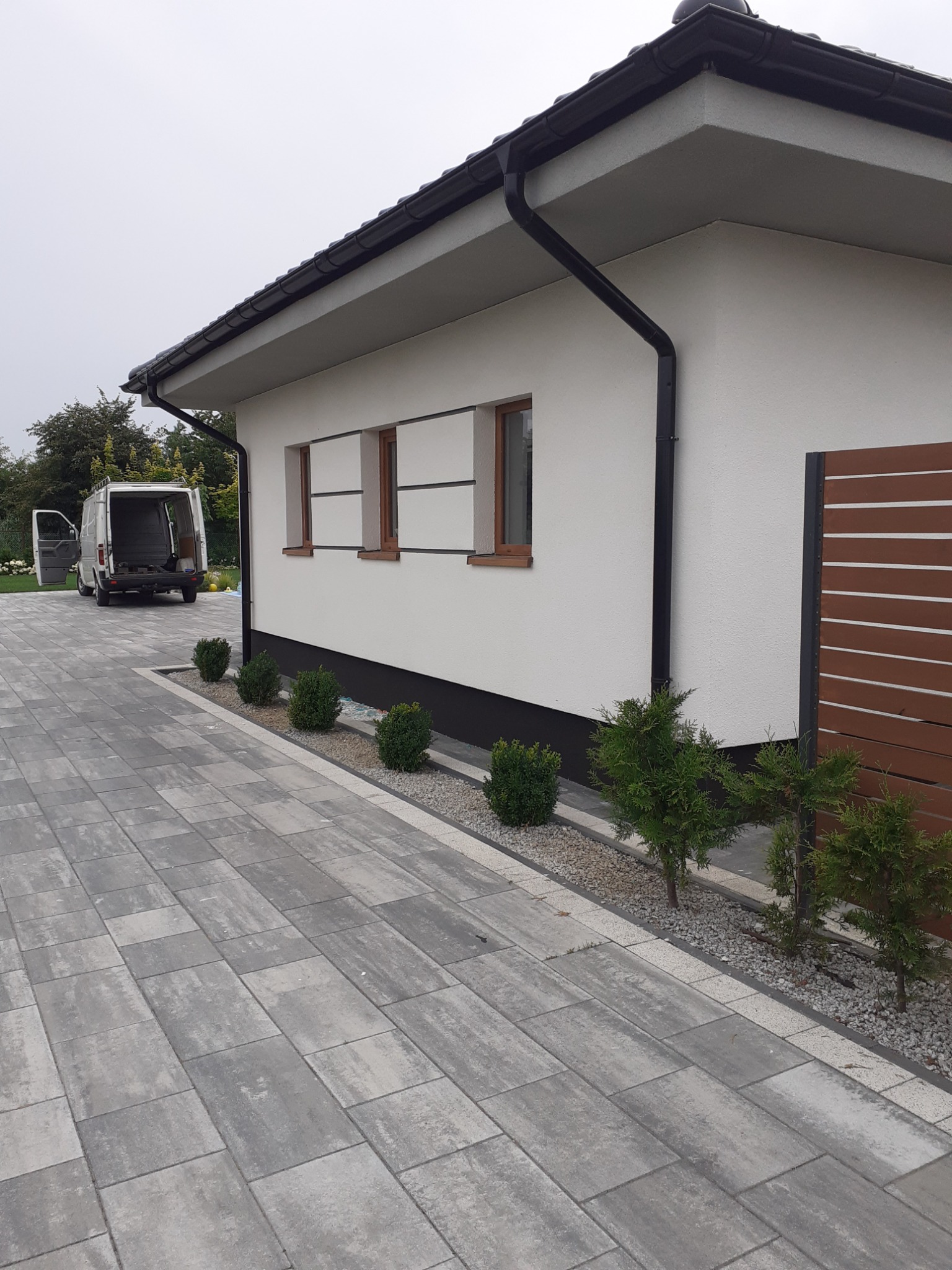 Biała elewacja domu jednorodzinnego z trzema oknami, czarną rynną i brązowym ogrodzeniem, widoczny szary podjazd z kostki brukowej i otwarty biały bus dostawczy w tle.