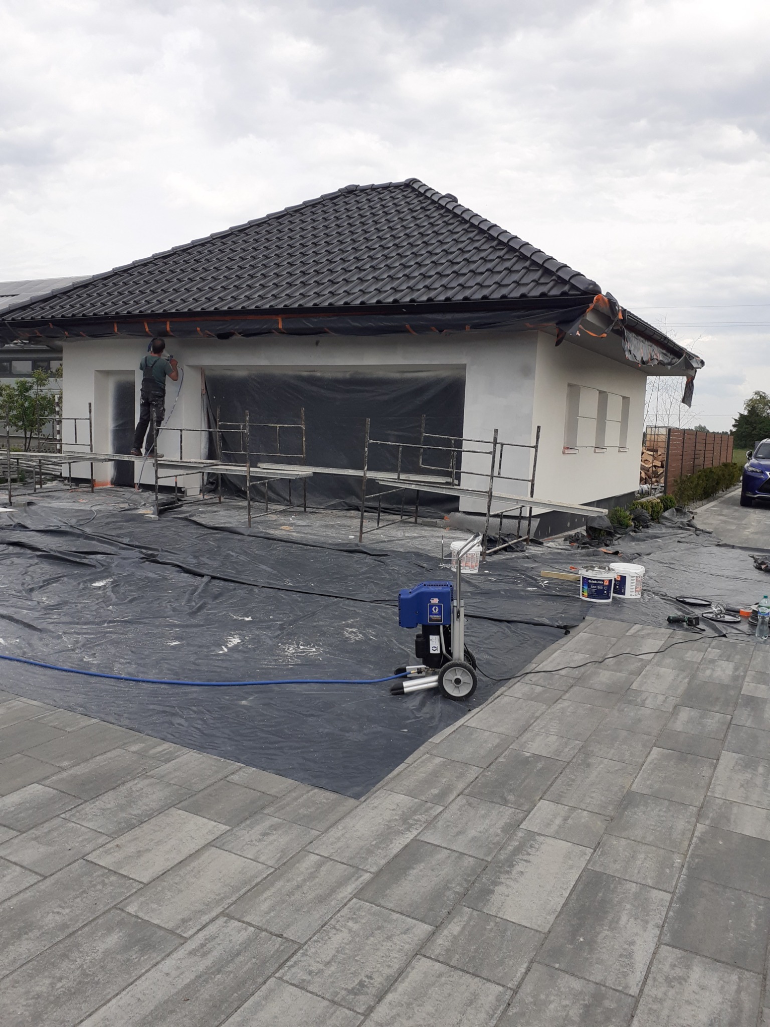 Pracownik na rusztowaniu maluje elewację domu natryskowo za pomocą agregatu malarskiego; zabezpieczona folią okna i teren wokół budynku.